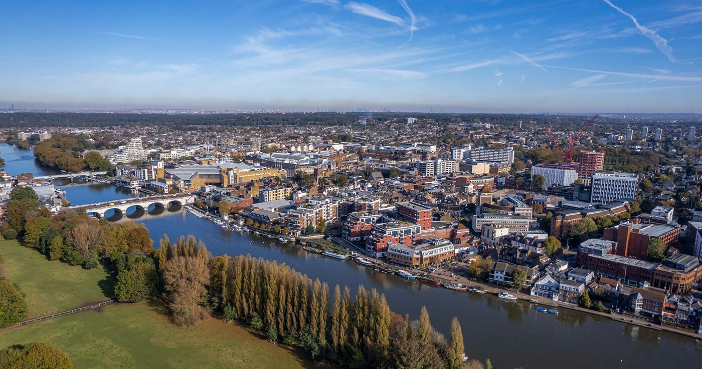Aerial view of a town along a river, bridge in the distance, buildings and trees, clear blue sky.