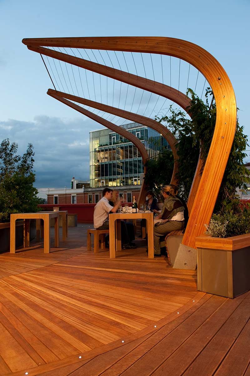 Rooftop restaurant with wooden canopy, people dining, evening setting with city buildings in the background.