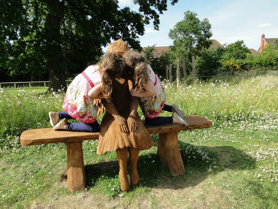 Two girls leaning over a wooden statue on a bench in a grassy park.