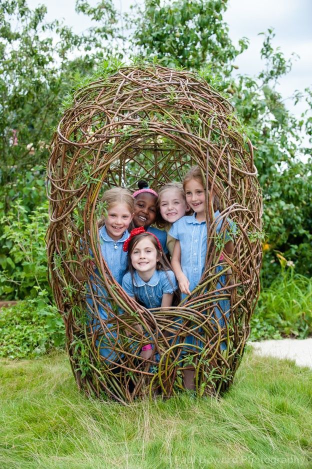 Five smiling children pose inside a large woven nest in a grassy garden.