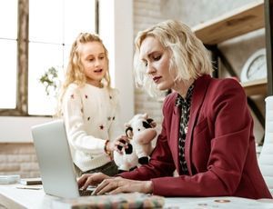 Child showing stuffed cow to mother in red blazer works while she works on a laptop.