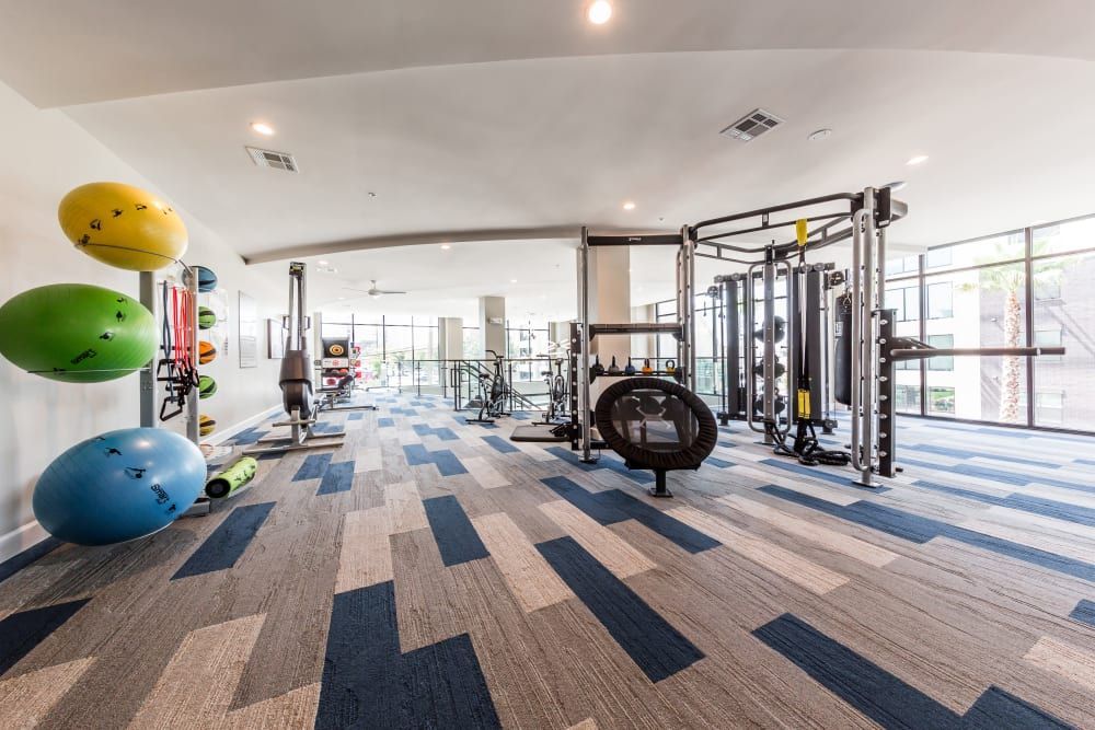 Image of a well-lit, modern gym interior with various exercise equipment. On the left, there's a rack of colorful stability balls, a rowing machine, and fitness mats. The center and right of the room feature a multi-station cable machine, free weights, and benches. Large windows provide natural light, and the room has a patterned blue and gray carpeted floor at Marq 31 in Houston, TX.