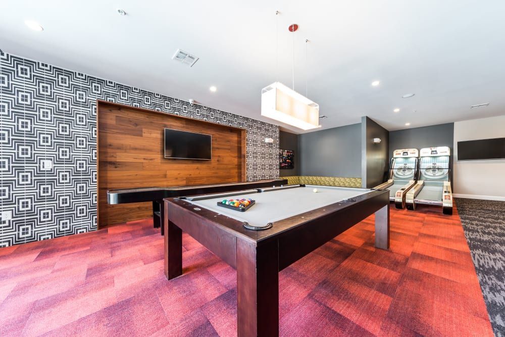 Spacious modern game room with geometric pattern wallpaper, a pool table in the foreground with balls racked for a game, a wall-mounted flat-screen TV, two bowling arcade machines at the back, and a sleek rectangular pendant light hanging overhead. The room features a richly textured red and gray carpet, and a wood-paneled wall adding warmth to the space at Marq 31 in Houston, TX.