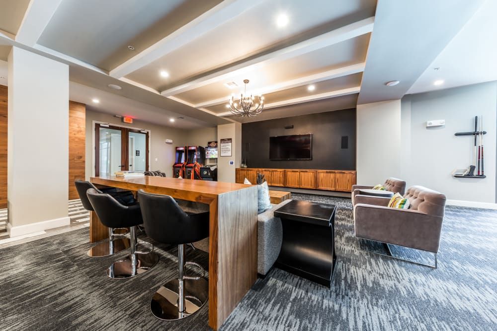 Modern and cozy communal lounge with a wooden bar counter and black bar stools, a seating area with plush sofas and colorful accent pillows, a large wall-mounted TV, and arcade game machines in the background. The space features wood paneling, stylish lighting fixtures, and a patterned carpet floor at Marq 31 in Houston, TX.