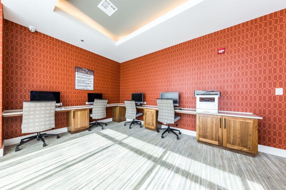 Modern business lounge with red patterned wallpaper, featuring a row of wooden desks with desktop computers and ergonomic office chairs, gray striped carpet flooring, and a sign on the central wall above the computers at Marq 31 in Houston, TX.