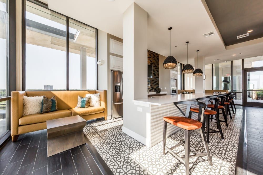 A modern, well-lit kitchen and seating area in a clubhouse with dark hardwood floors and large windows offering an expansive view. A mustard yellow sofa with decorative pillows sits adjacent to the windows, accompanied by a unique wooden coffee table. The kitchen features a long white island with a sleek surface, bar seating with stylish black stools with red cushioning, and pendant lighting with black and gold patterned shades. Stainless steel appliances and a patterned backsplash add to the contemporary aesthetic at Marq 31 in Houston, TX.
