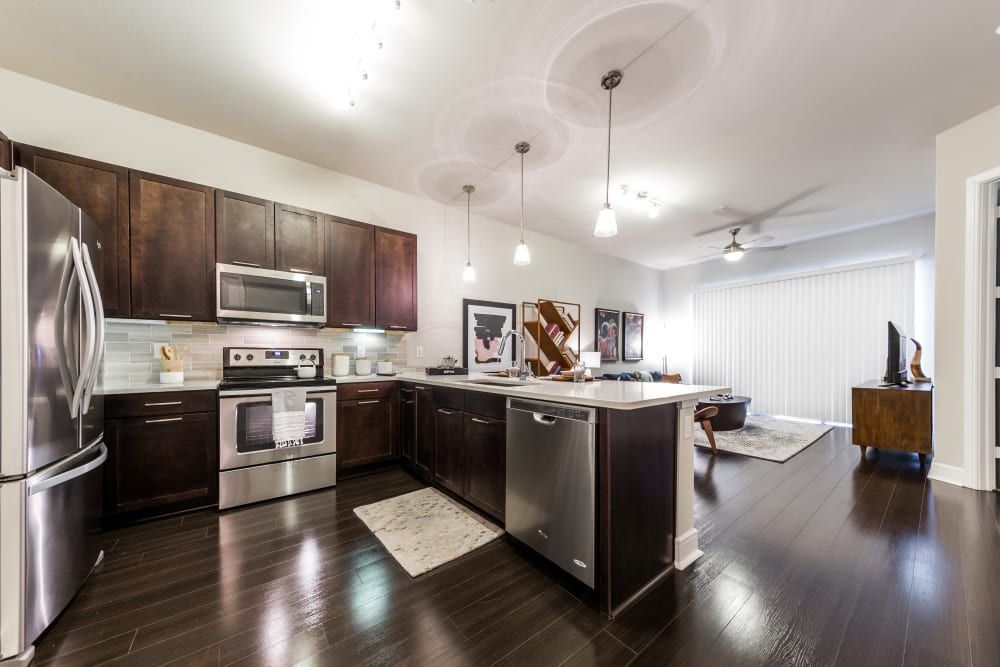 Modern kitchen with dark wood cabinetry and stainless steel appliances, including a refrigerator, microwave, and oven. An island with a sink and dishwasher is at the center, leading to an open-plan living area with a white couch and a desk with a computer at Marq 31 in Houston, TX.