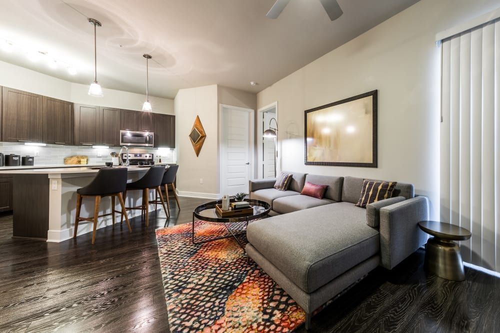 Modern open-plan living room connected to a kitchen, featuring dark wood flooring, a gray sectional sofa adorned with colorful cushions, and a geometric-patterned area rug. The kitchen includes dark wood cabinets, stainless steel appliances, white countertops, and a breakfast bar with black bar stools. Ambient lighting is provided by pendant lights over the kitchen counter and natural light coming through vertical blinds at Marq 31 in Houston, TX.