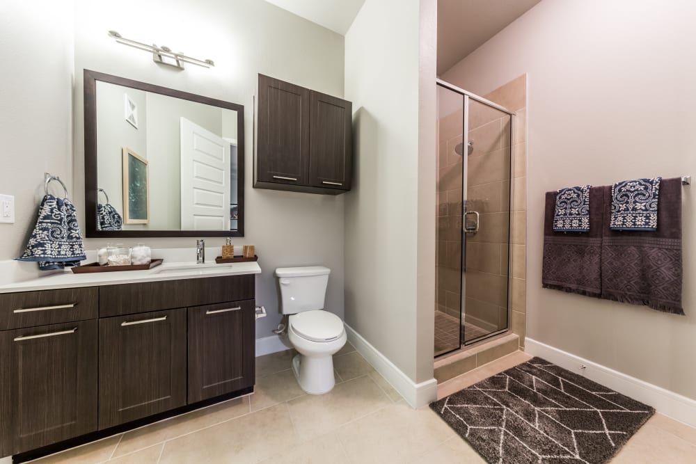 Modern bathroom interior with dark wood cabinetry, beige tiling, and glass shower door. A large mirror is above a vanity with a white countertop, and two patterned blue towels hang on the wall beside the shower. The floor is adorned with a geometric-patterned rug at Marq 31 in Houston, TX.