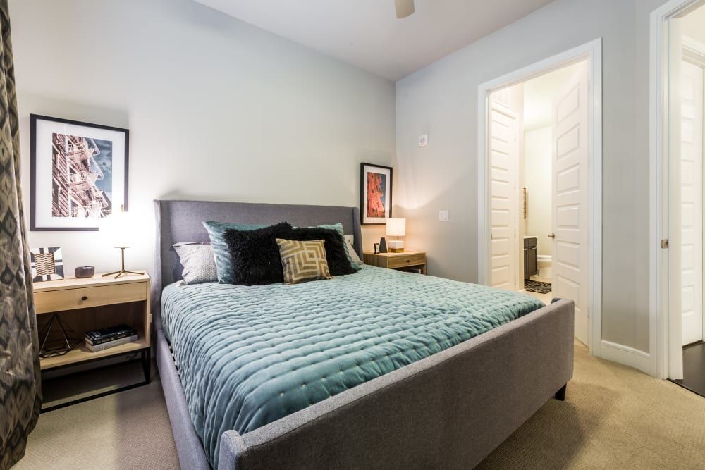 A cozy bedroom with a large gray upholstered bed, teal bedding, mixed patterned pillows, and two wooden nightstands with lamps. Wall art hangs above each nightstand, and the room features a neutral color palette with white doors leading to another room or closet at Marq 31 in Houston, TX.