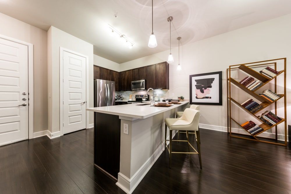 Modern kitchen interior with dark hardwood floors, dark brown cabinets, and white countertops. Features stainless steel appliances, a kitchen island with a white chair, pendant lights, a geometric bookshelf with books, and a framed artwork on the wall at Marq 31 in Houston, TX.