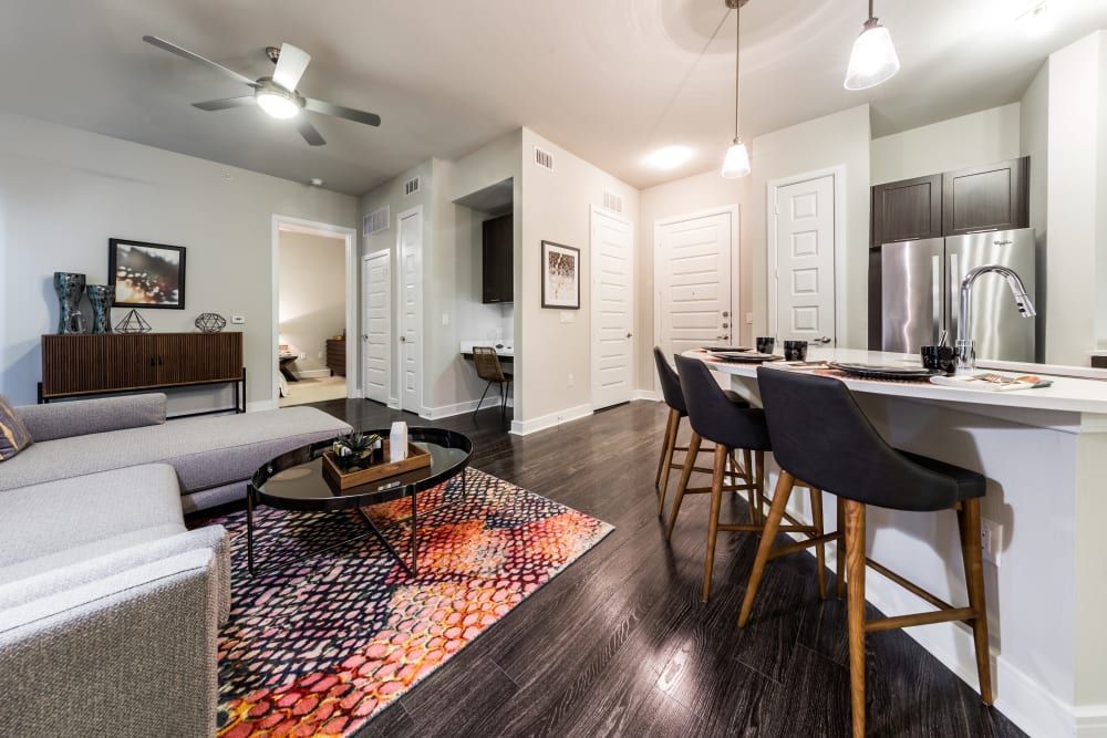 Modern open-plan living room and kitchen with bar stools at a counter, a colorful area rug, dark hardwood floors, and clean, minimalist decor at Marq 31 in Houston, TX.