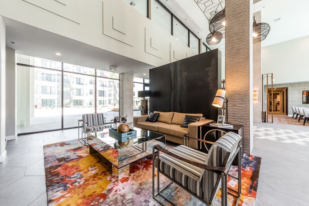 Modern and spacious lobby interior featuring a comfortable seating area with striped armchairs, brown leather sofa, and glass coffee tables on a vibrant multicolored rug. High ceilings with unique geometric lighting fixtures, large windows allowing natural light, and decorative elements create a welcoming and stylish atmosphere at Marq 31 in Houston, TX.