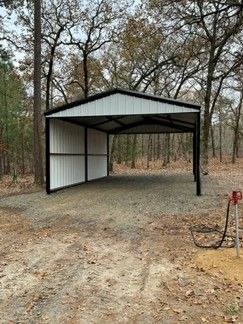 White and black metal carport on gravel in a wooded area.