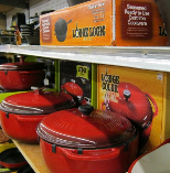 Red ceramic grills on a store shelf beside orange packaging and a black kettle grill case