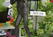 Decorative dark statue beside a white sign reading “SOLAR,” surrounded by green plants and colorful flowers