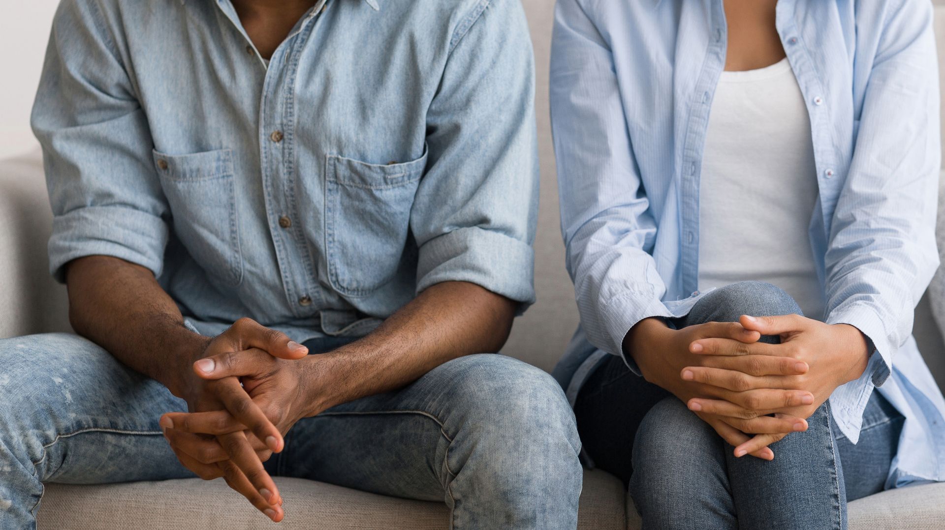 Two people sit on a couch, hands clasped. Both wear blue button-down shirts and jeans.
