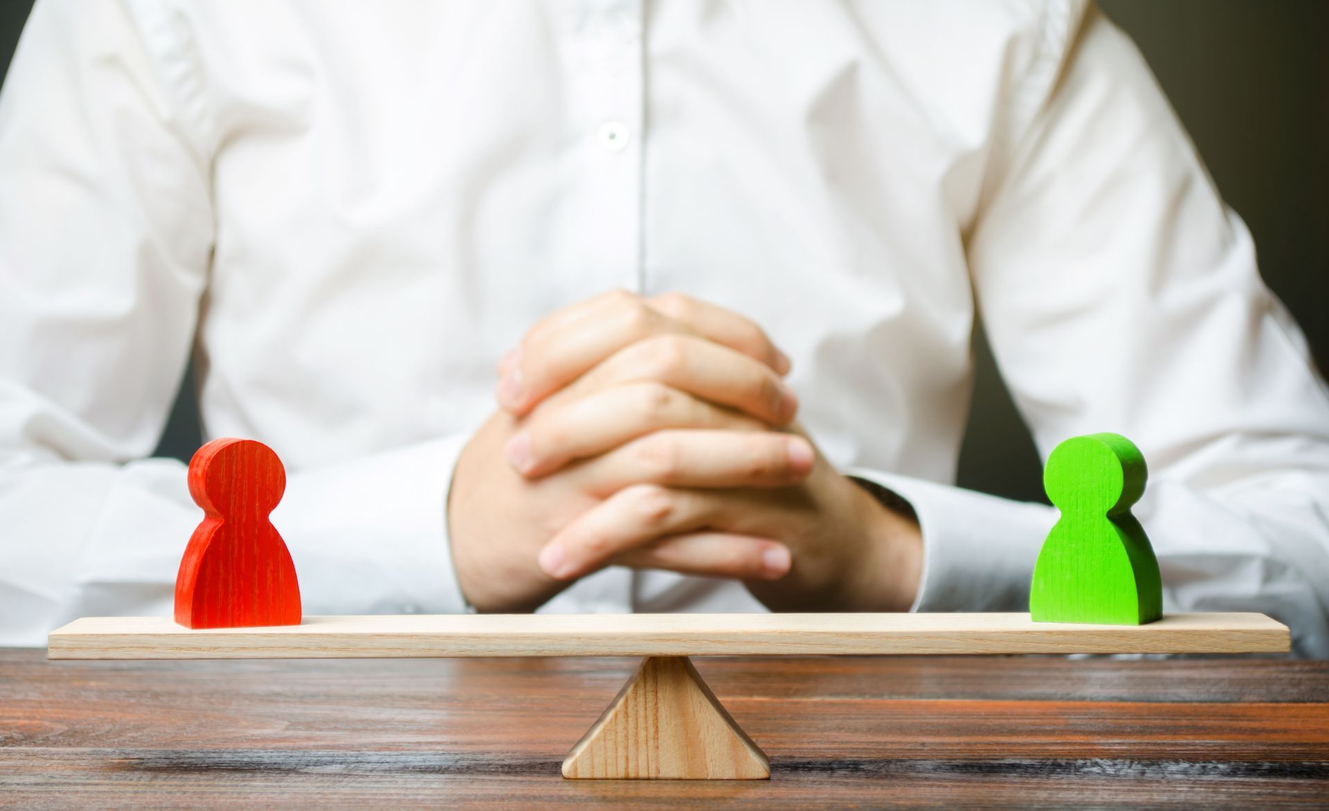 Red and green wooden figures balanced on a small scale, with a person sitting behind them with folded hands.