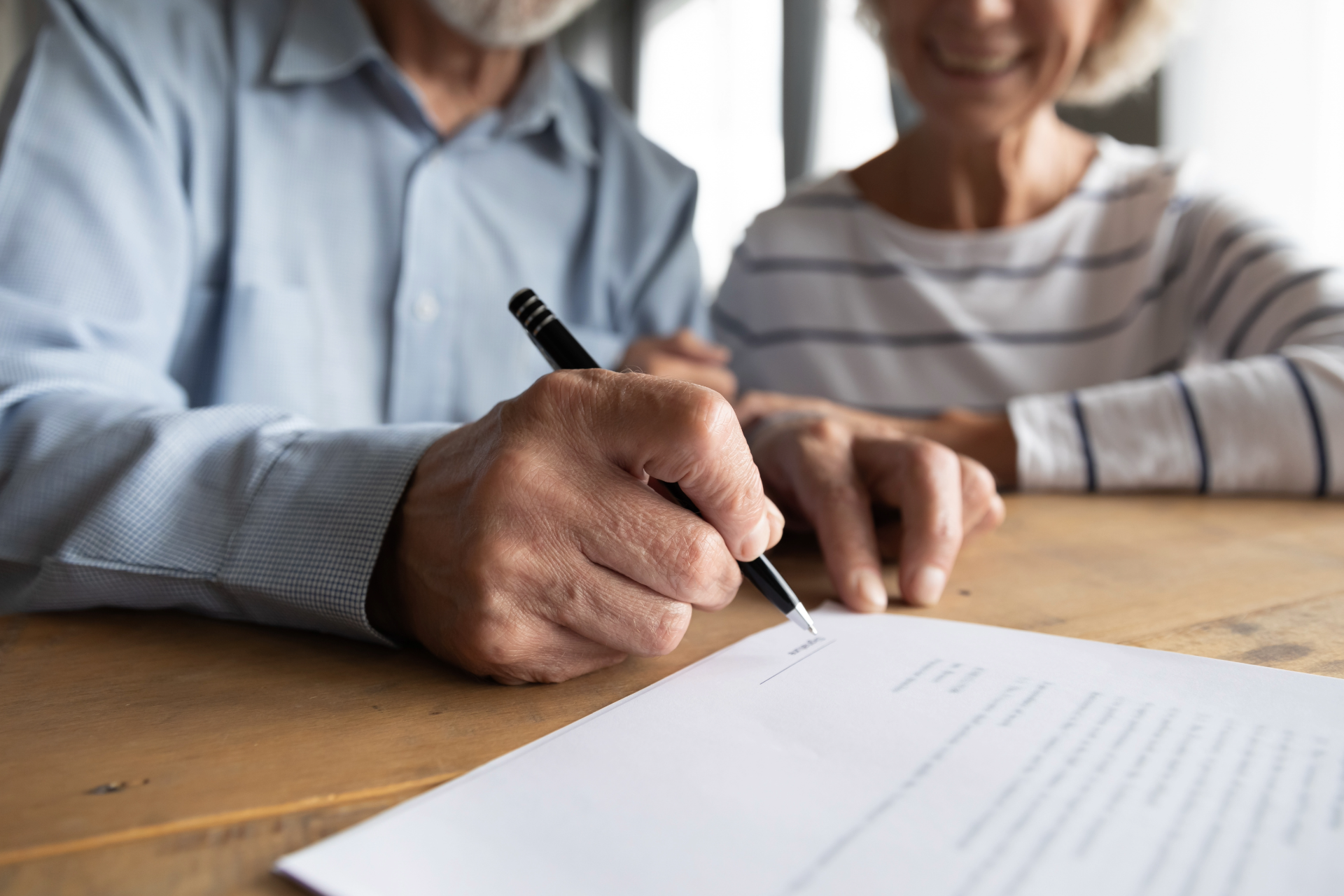 Older person signing document at a table, another person smiling in the background.