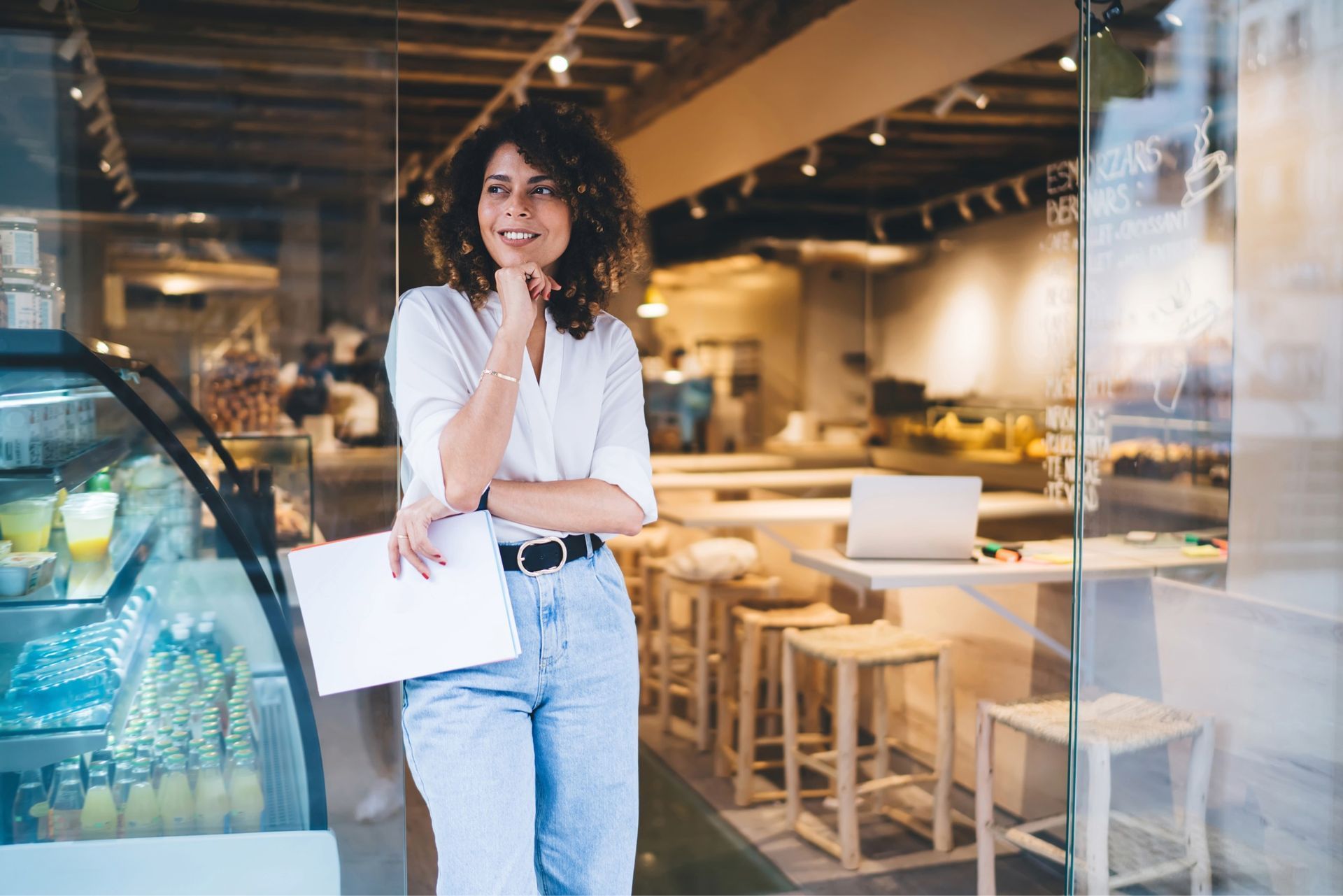 Woman leaning in doorway of cafe, holding papers, looking thoughtful.