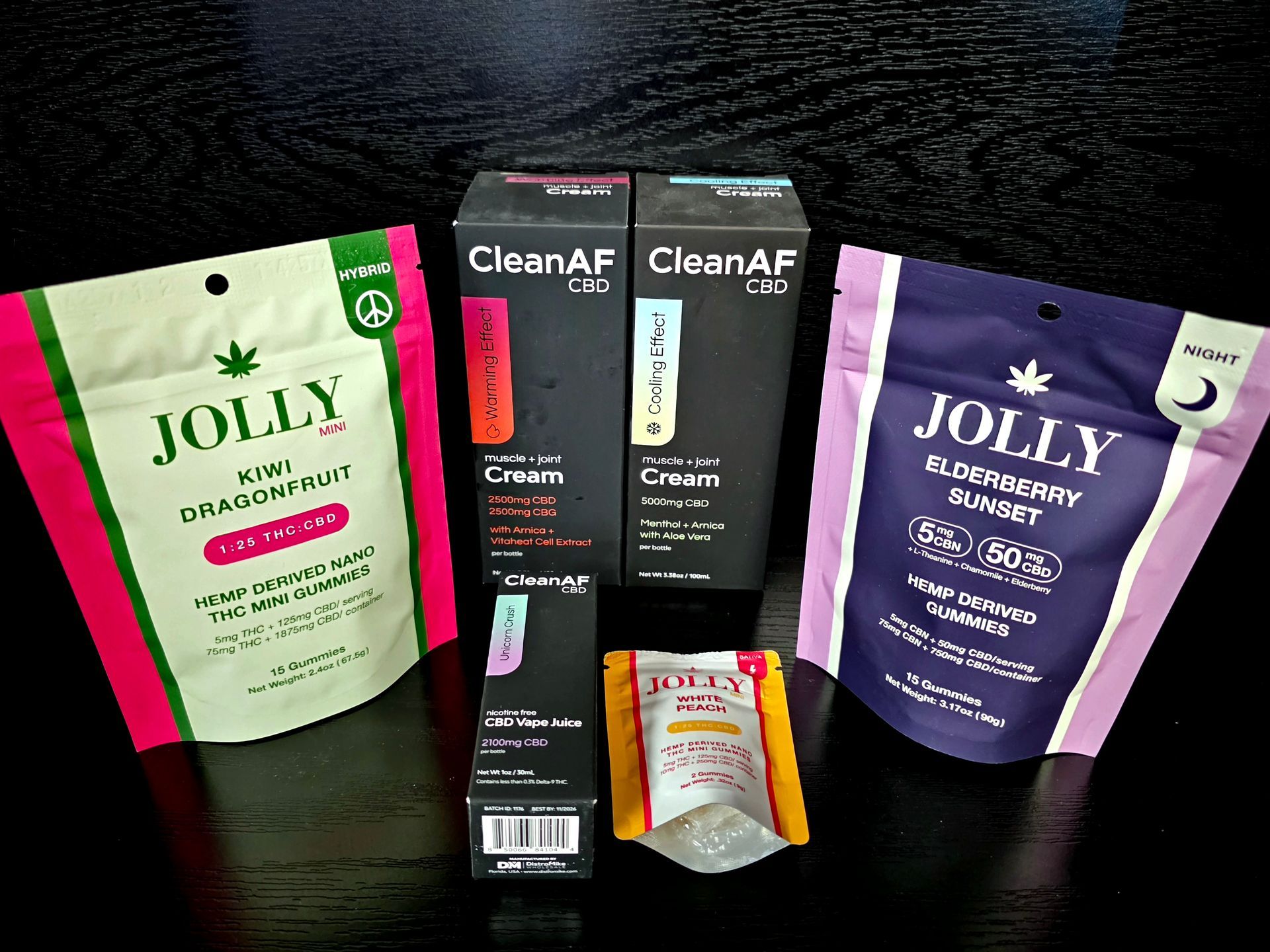 A collection of CleanAF and Jolly brand CBD products, including two cream boxes and two gummy pouches on a black surface.