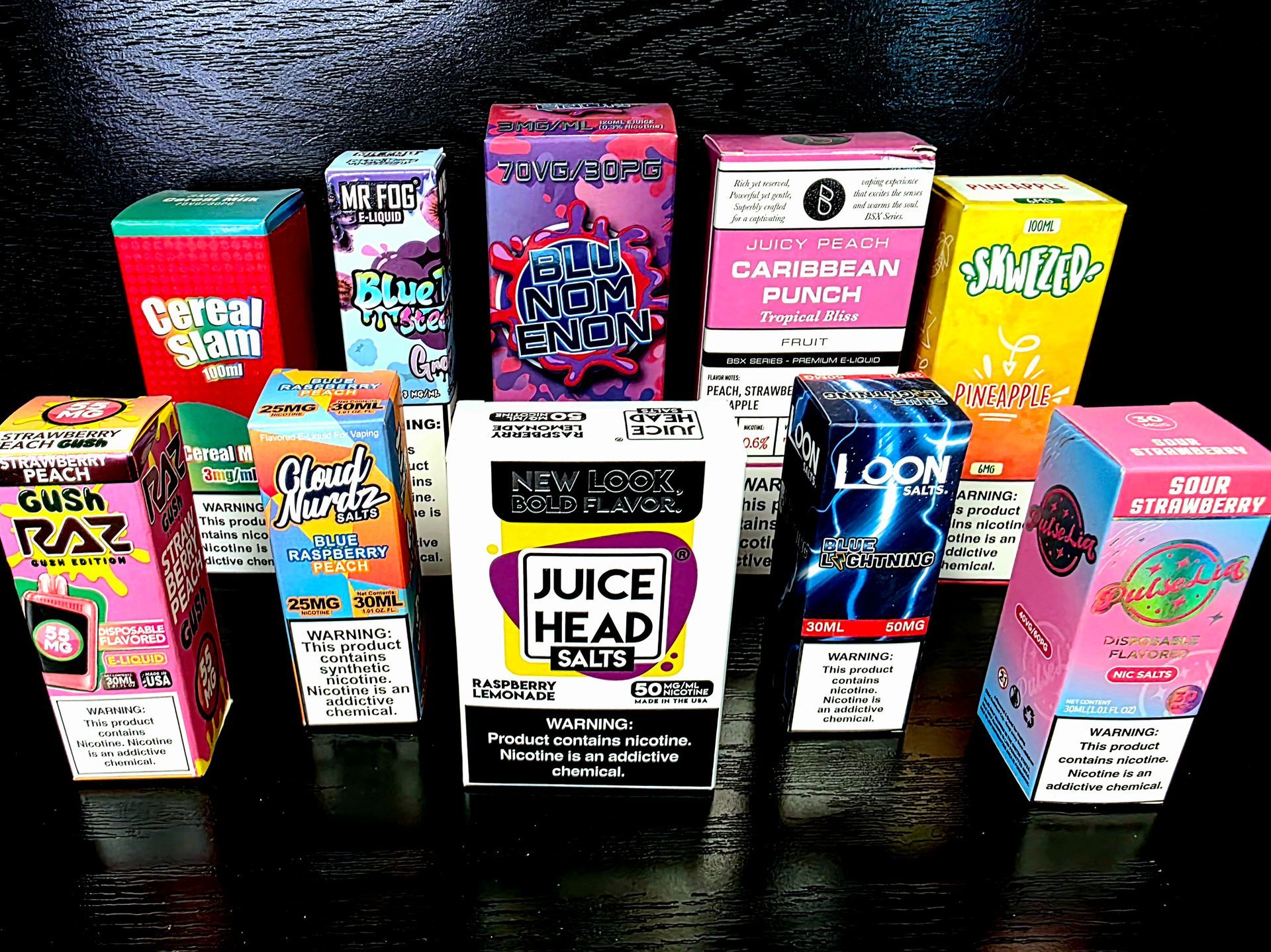 A display of various colorful e-liquid or vape juice boxes arranged on a dark, reflective surface.