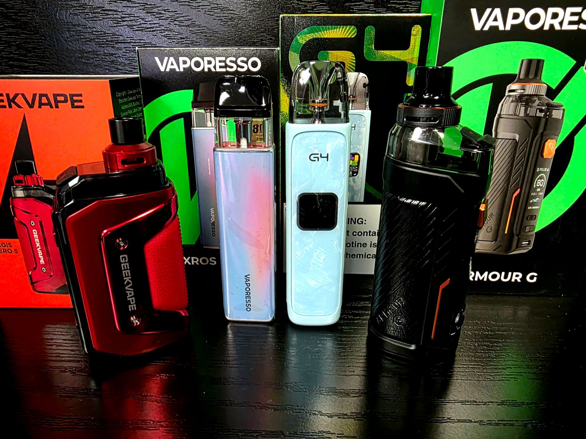 Four Vaporesso and Geekvape vaping devices are displayed in front of their respective retail boxes on a black surface.