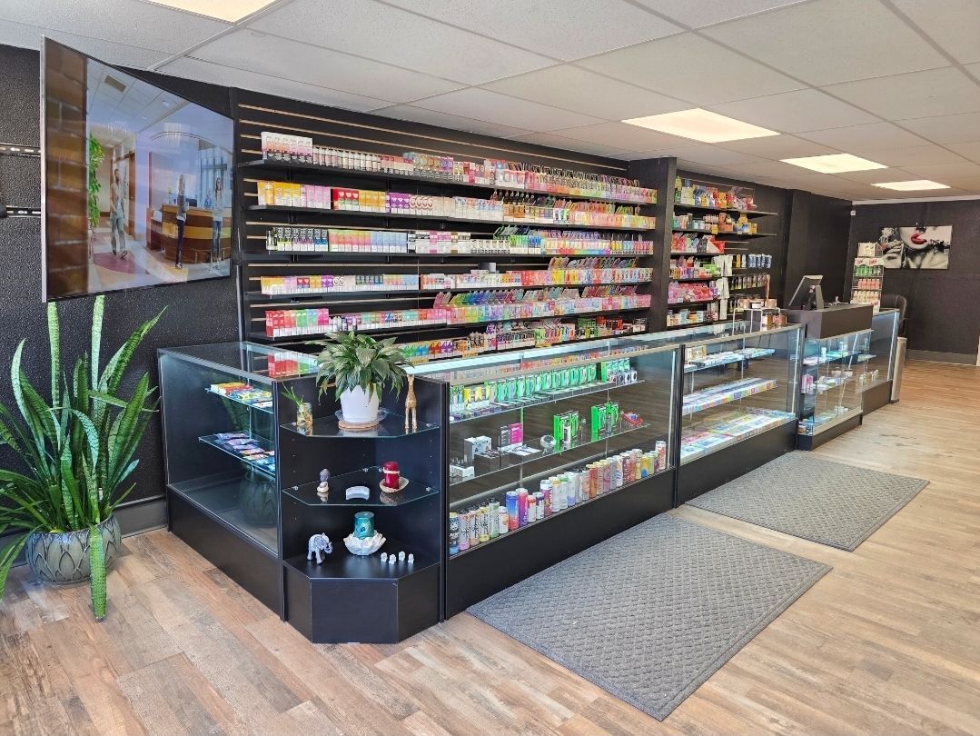 Interior view of a smoke shop featuring long glass display counters, shelves packed with products, and a potted plant.