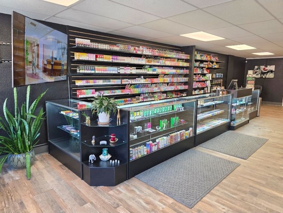 Interior view of a smoke shop featuring long glass display counters, shelves packed with products, and a potted plant.