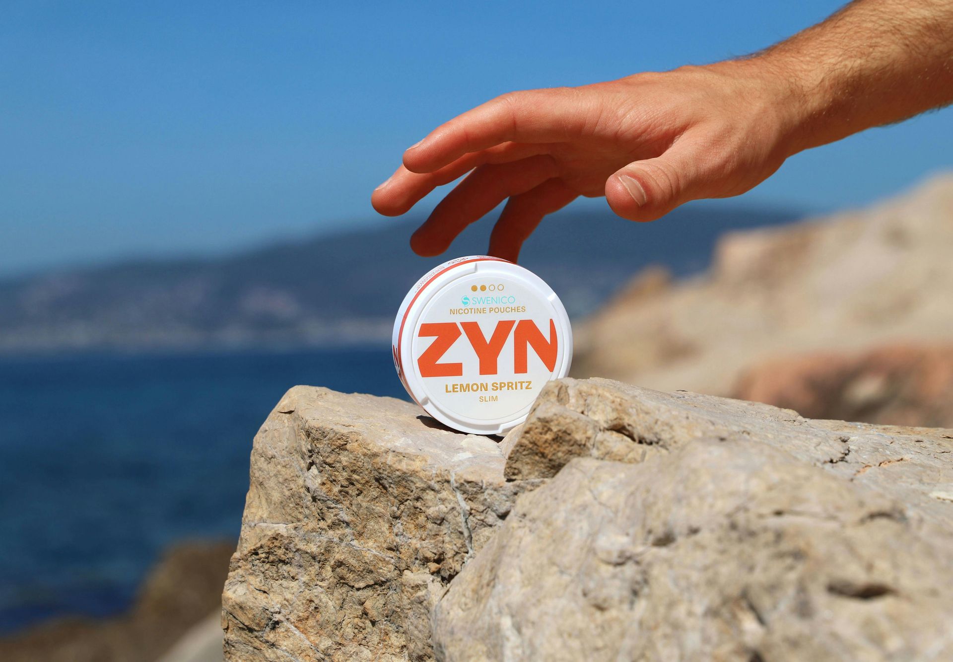 A hand reaches toward a ZYN nicotine pouch tin balanced on a rock overlooking the ocean.