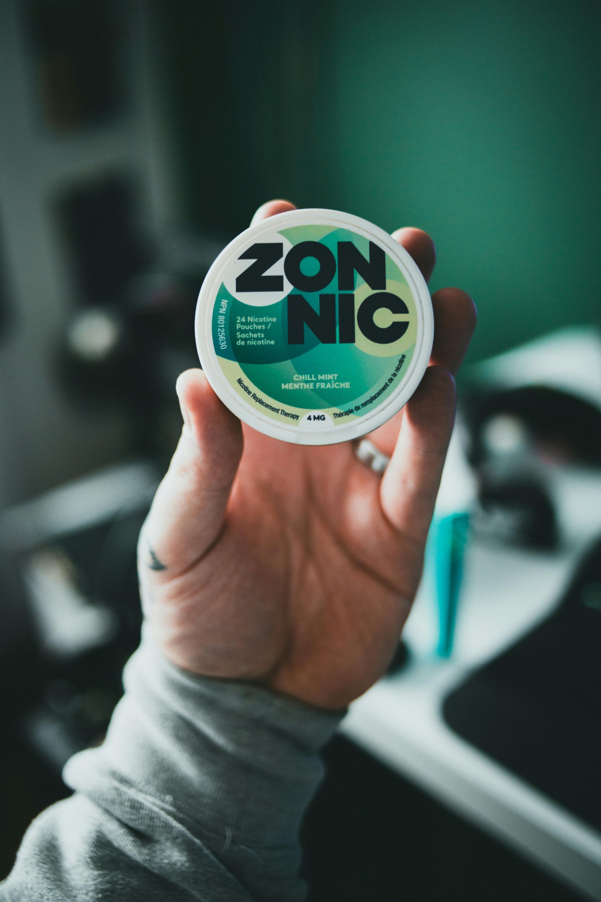 A hand holds a circular container of ZONNIC nicotine pouches, featuring a green and white design, against a blurred room.