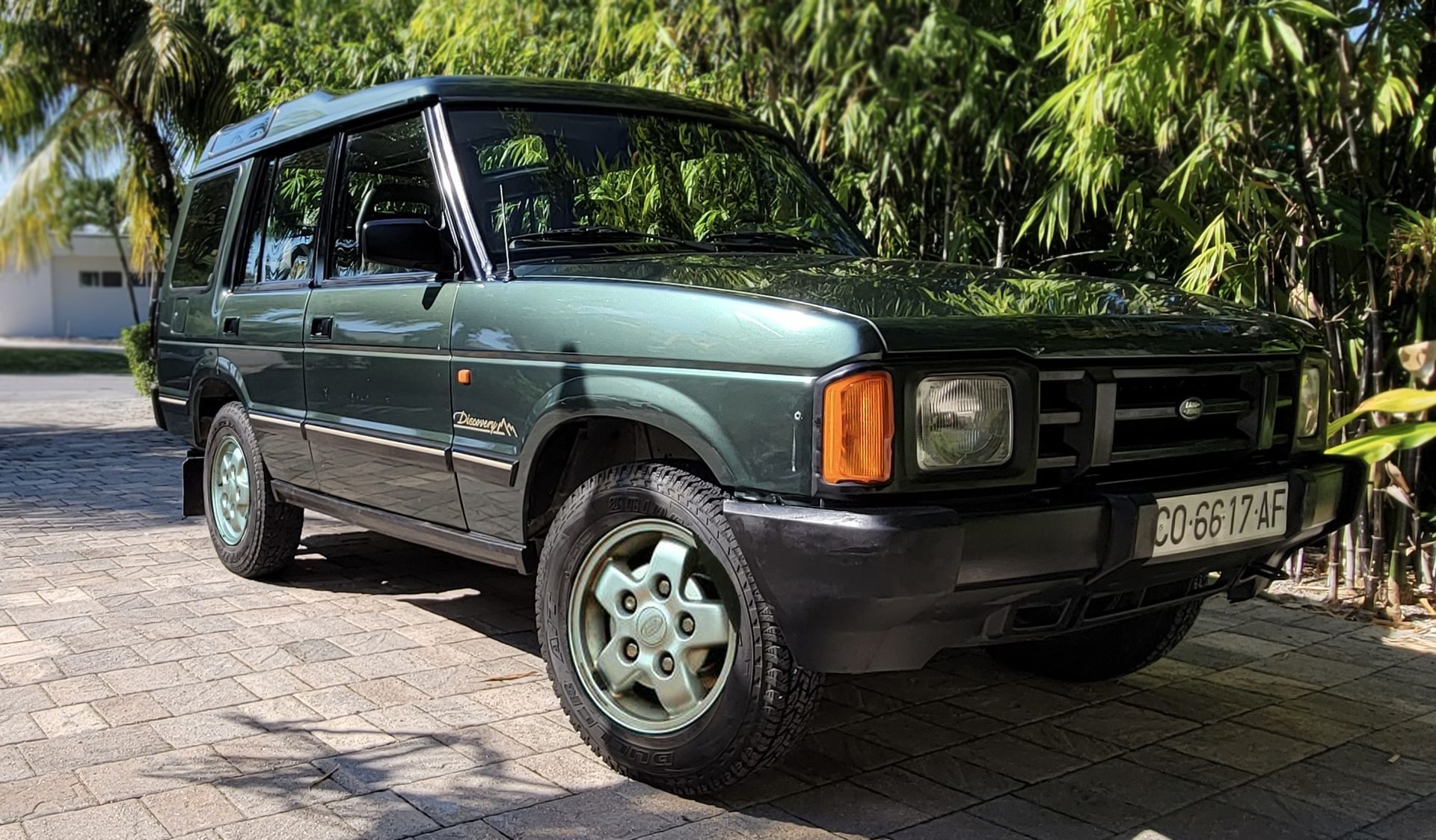 Green Land Rover Discovery SUV parked on a cobblestone driveway, surrounded by greenery.
