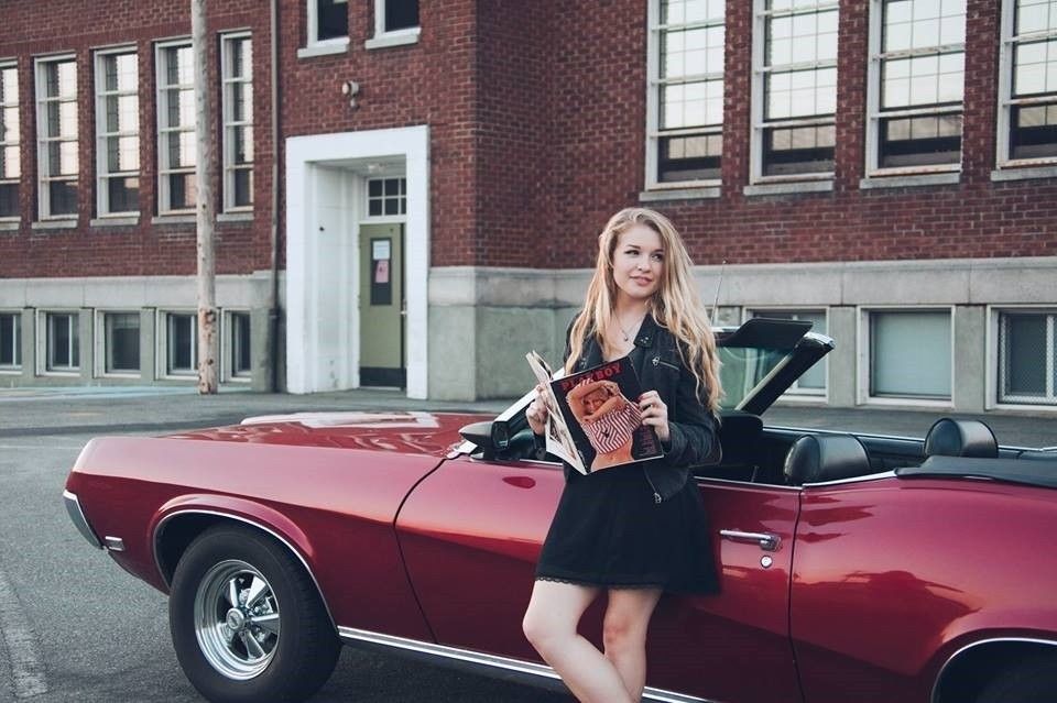 Woman leaning on red convertible, holding magazine, in front of a brick building.