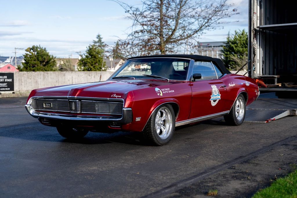 Woman leaning on red convertible, holding magazine, in front of a brick building.