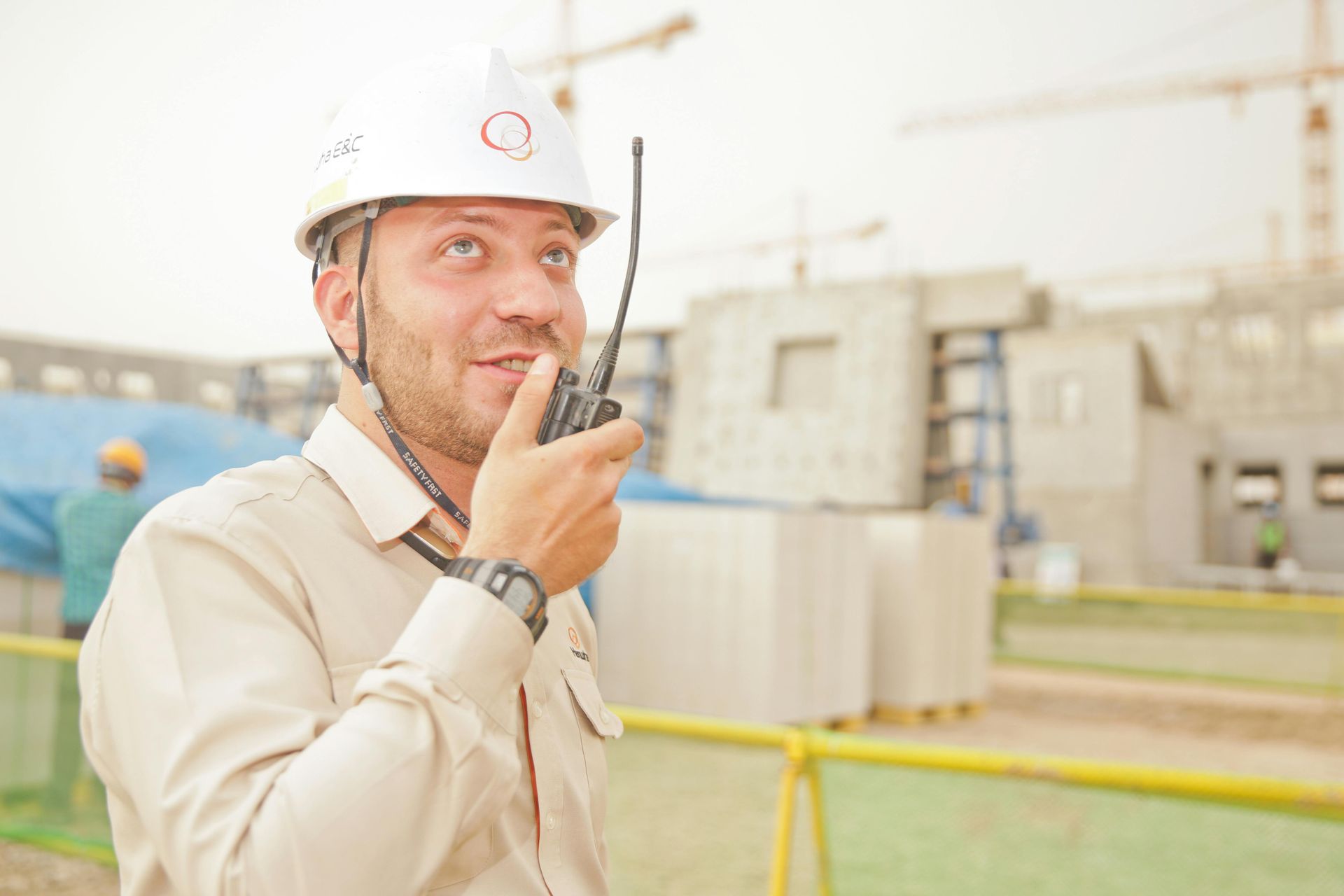 Construction worker in a white hard hat and tan shirt using a walkie-talkie at a construction site.
