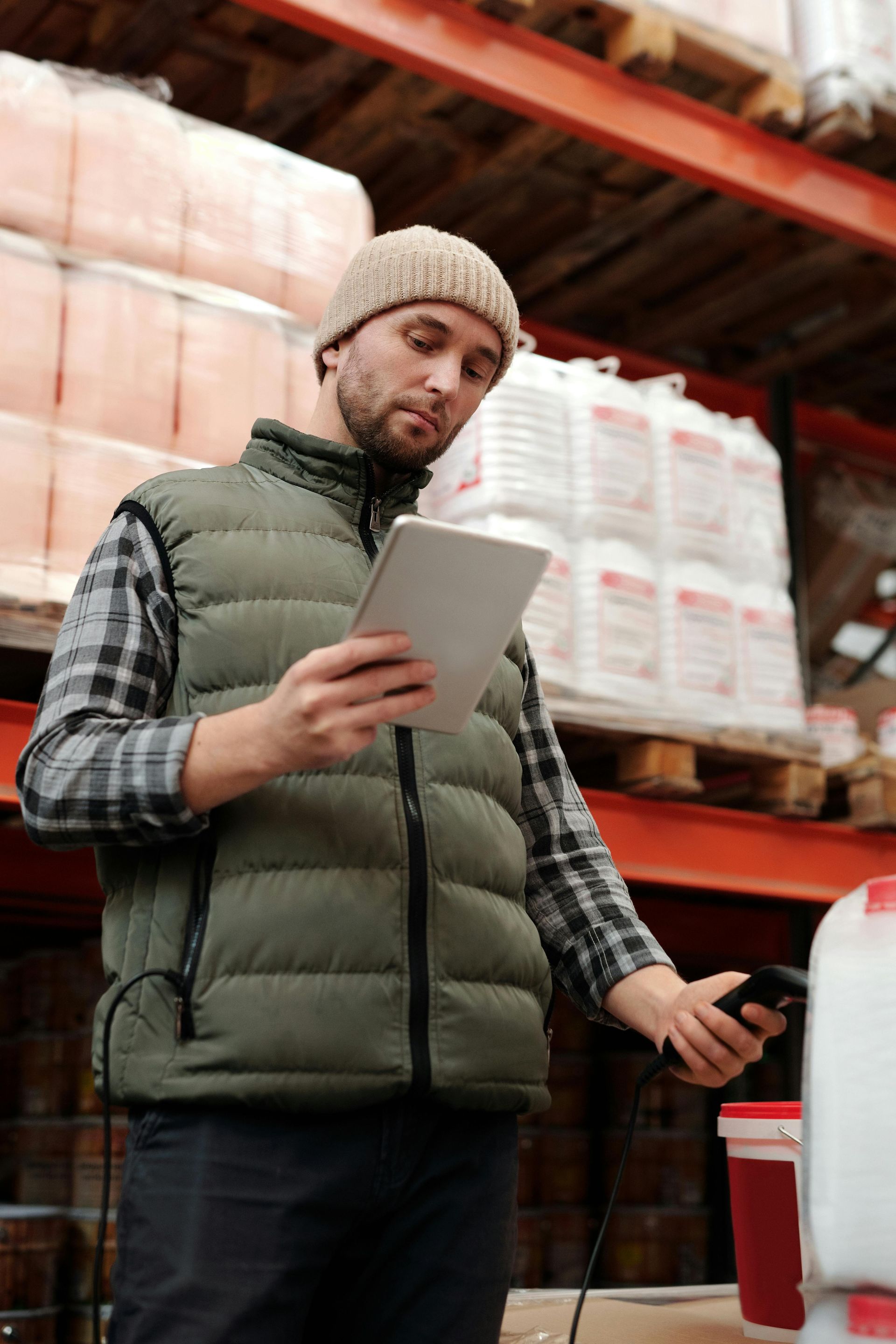Man in warehouse vest uses tablet and scanner near shelves of product.