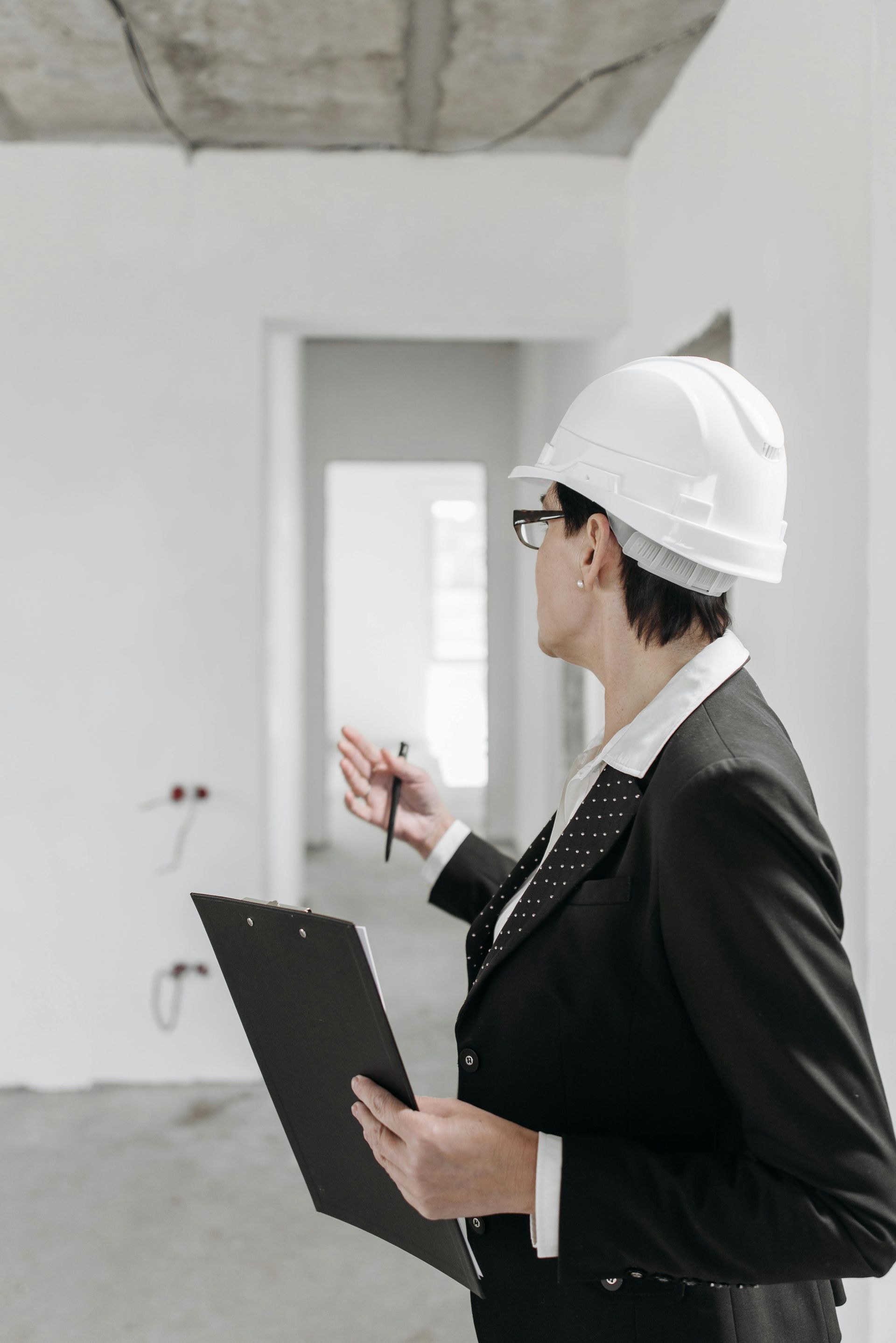 Woman in hard hat and suit inspecting a construction site, holding a clipboard and pen.