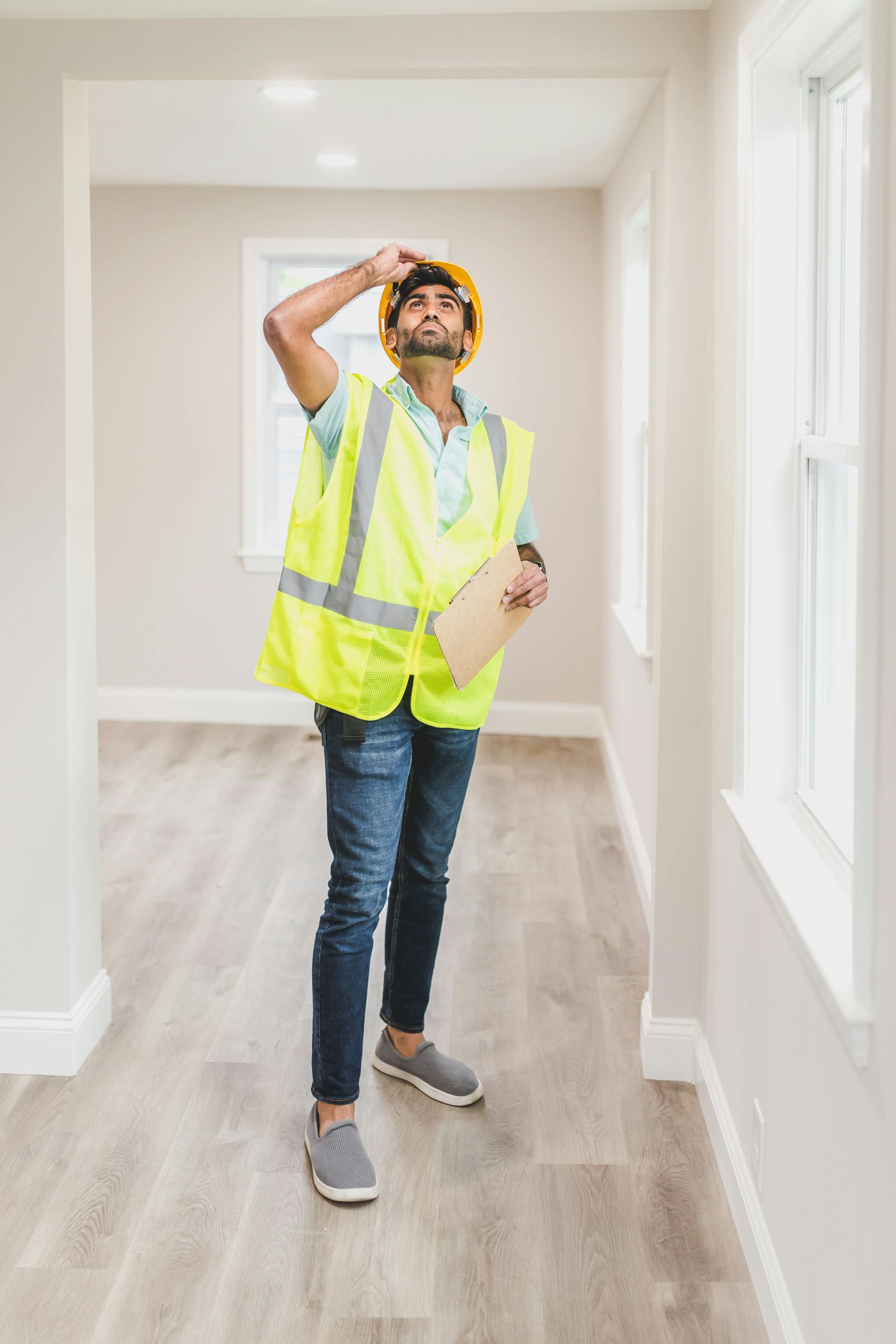 Construction worker in a yellow vest and hard hat inspects a hallway, holding a clipboard and looking upward.