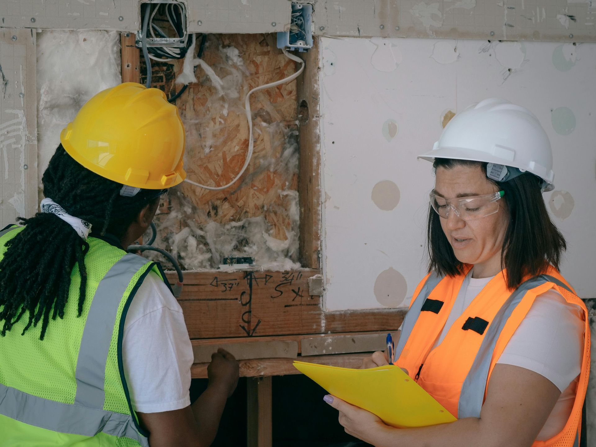 Two construction workers examining wall damage; one writing on clipboard.