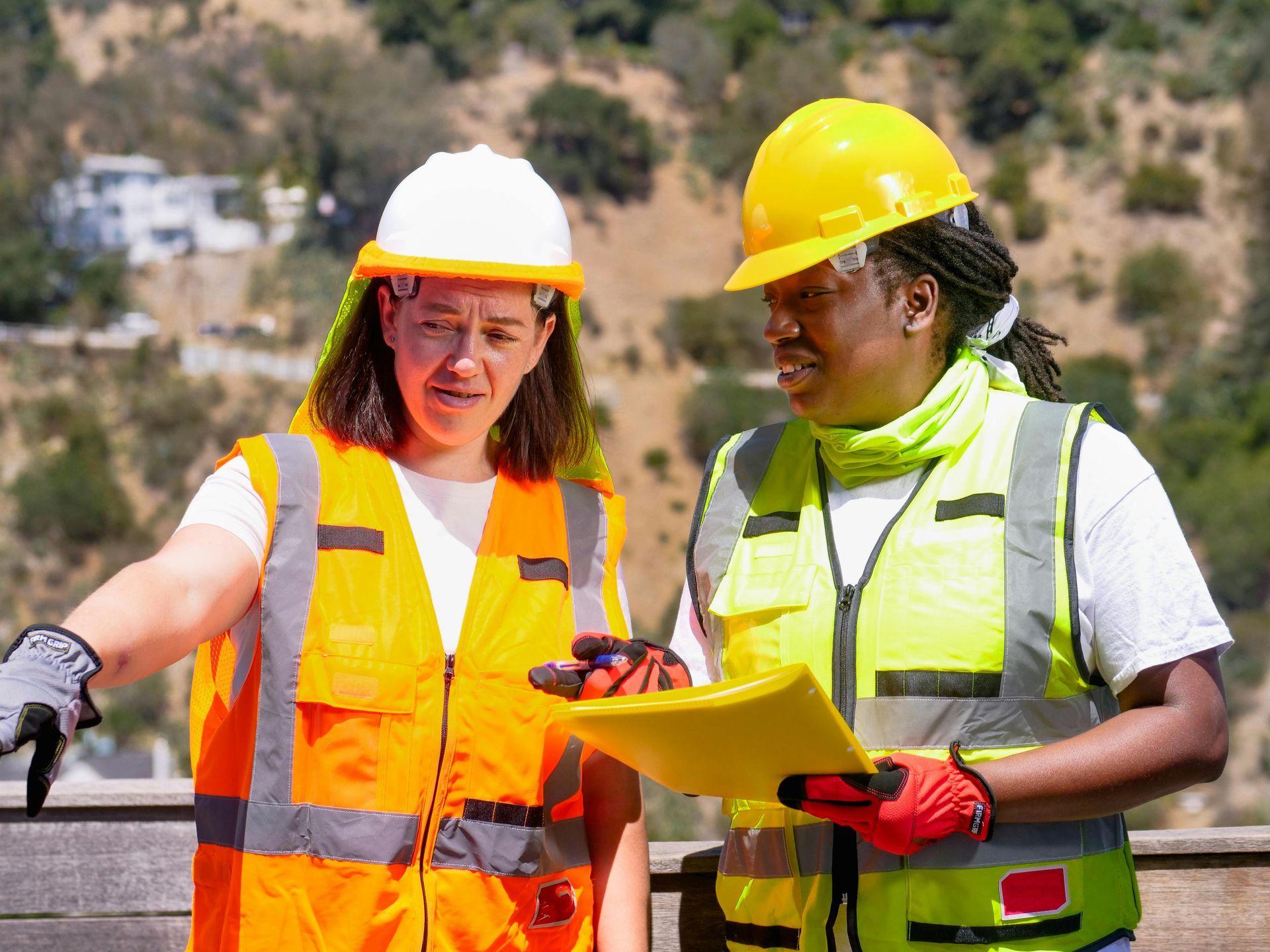 Two construction workers in safety vests and hard hats reviewing plans, outdoors.