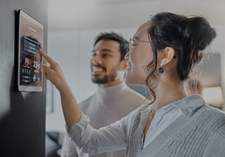 Woman touching a smart home control panel. A man smiles in the background. Indoor setting, white walls, daylight.