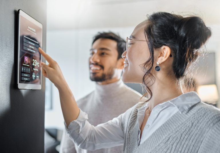 Woman adjusting smart home control on a tablet; man smiles in background. Bright interior setting.