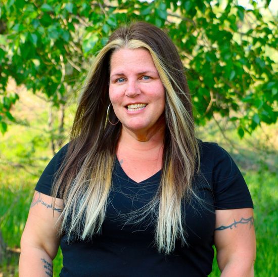 A woman is standing outside with trees and grass in the background.