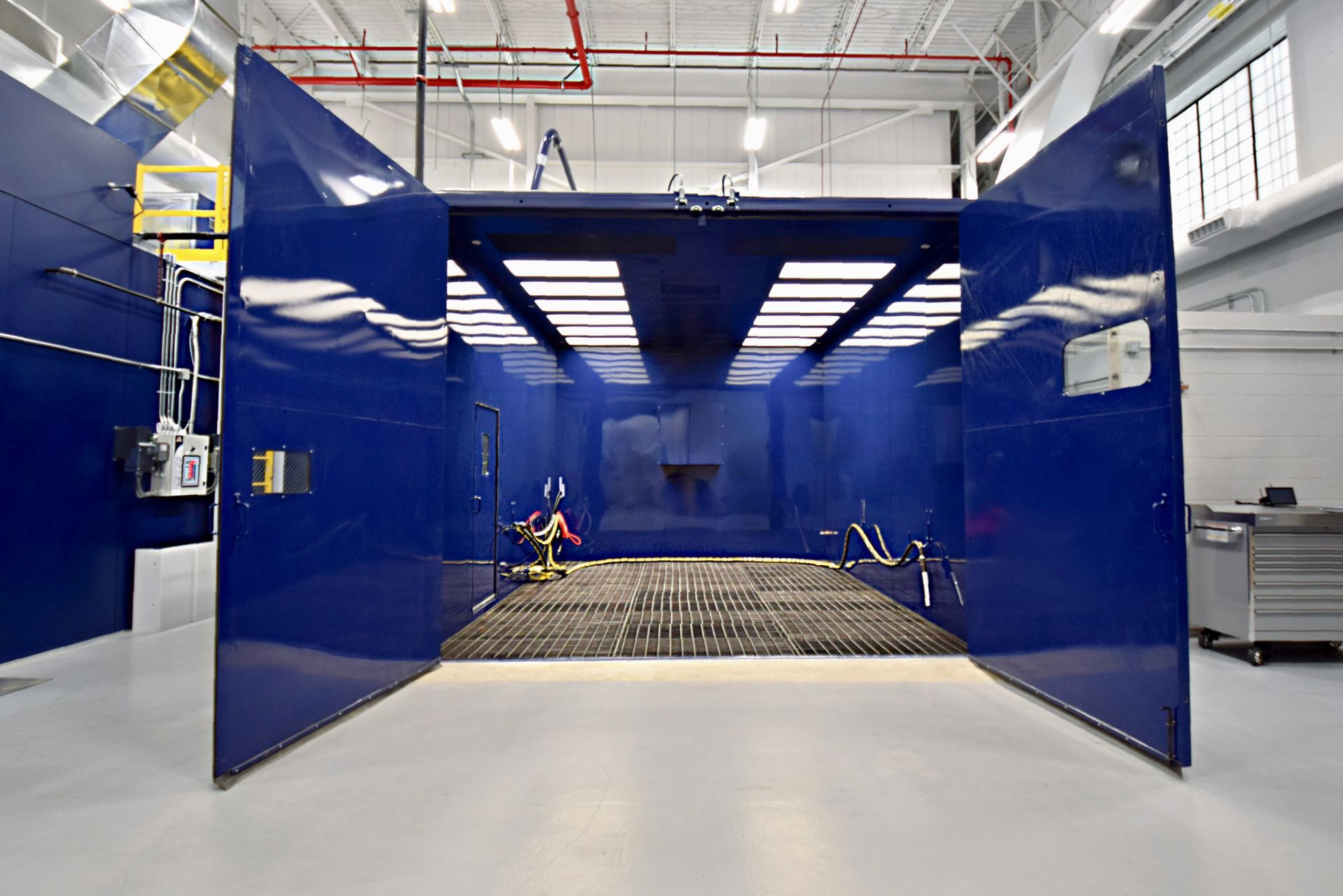 Blue industrial paint booth, open doors, gridded floor, bright interior lighting, white walls.