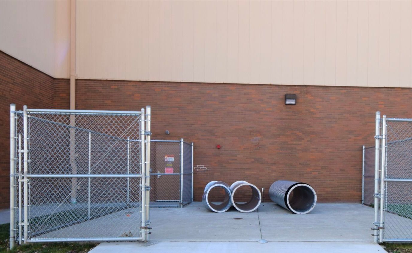 Chain link fence with open gates, brick wall, concrete ground, and three large pipes.