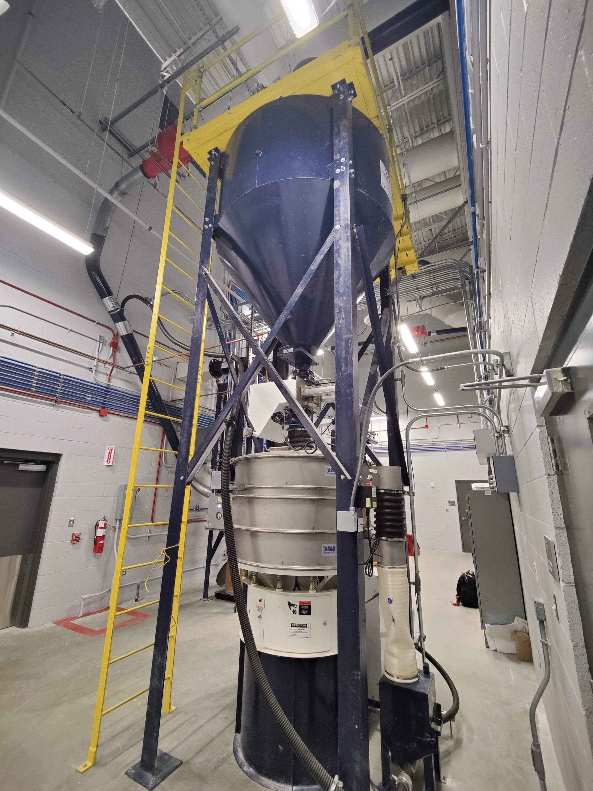 Tall industrial machinery in a factory setting. A blue conical container on a metal frame with a yellow ladder.