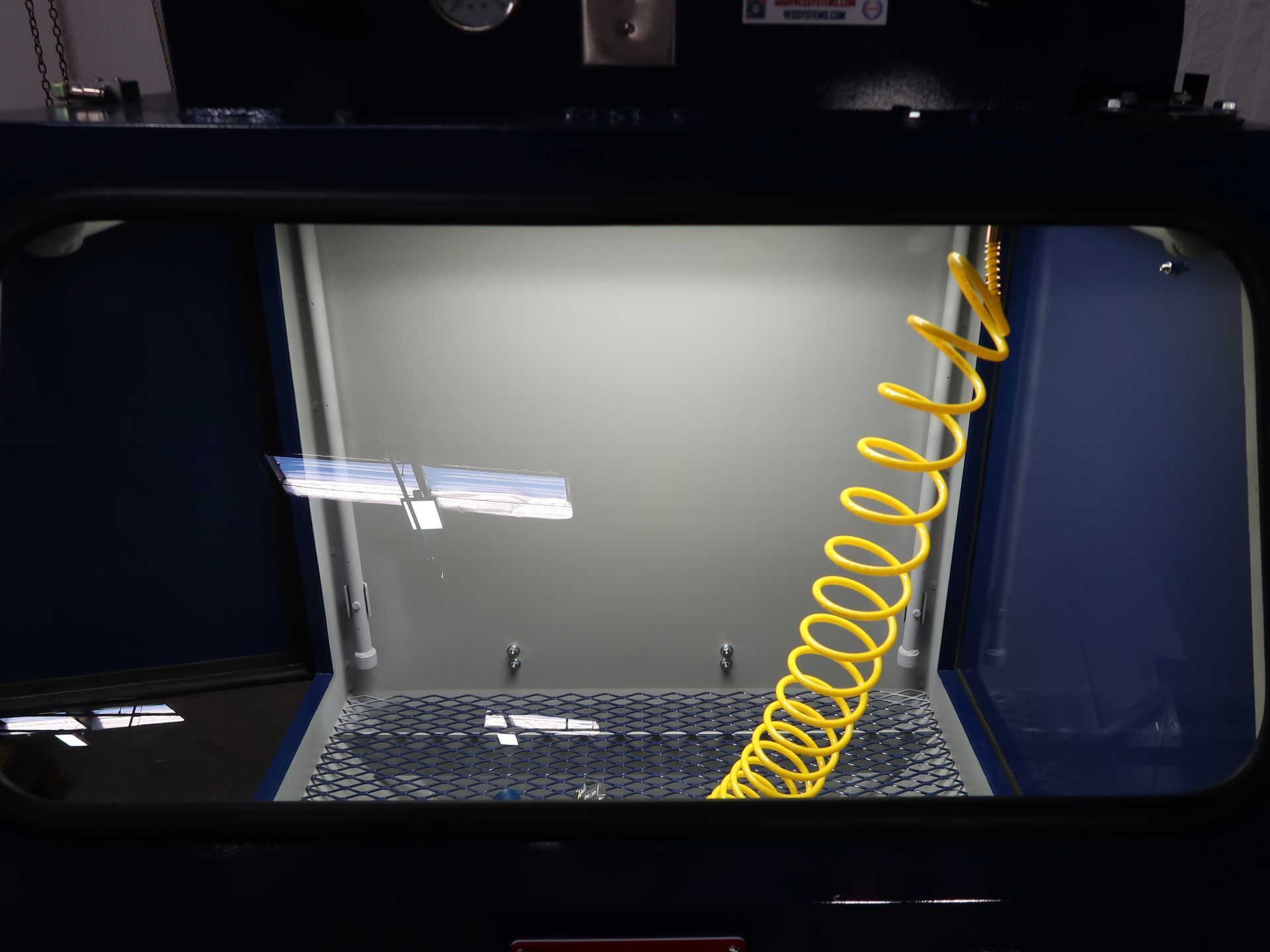 Inside a blue parts washer, a metal item is suspended. A yellow coiled hose is visible.