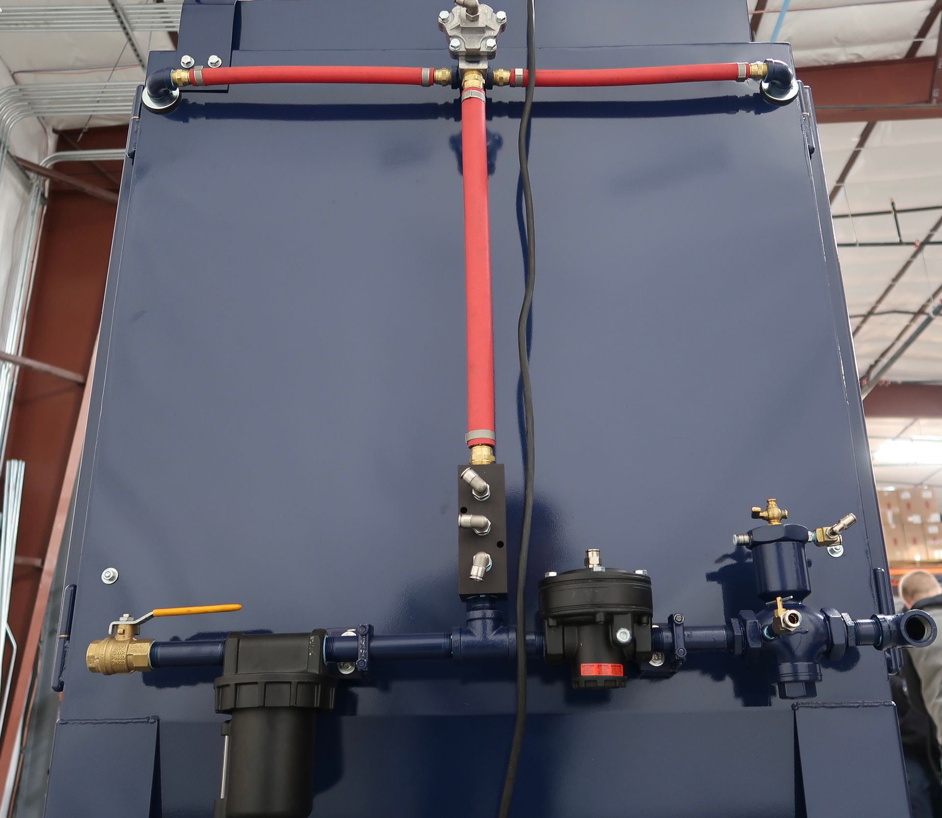 Blue industrial equipment with red and blue pipes and valves against a warehouse wall.