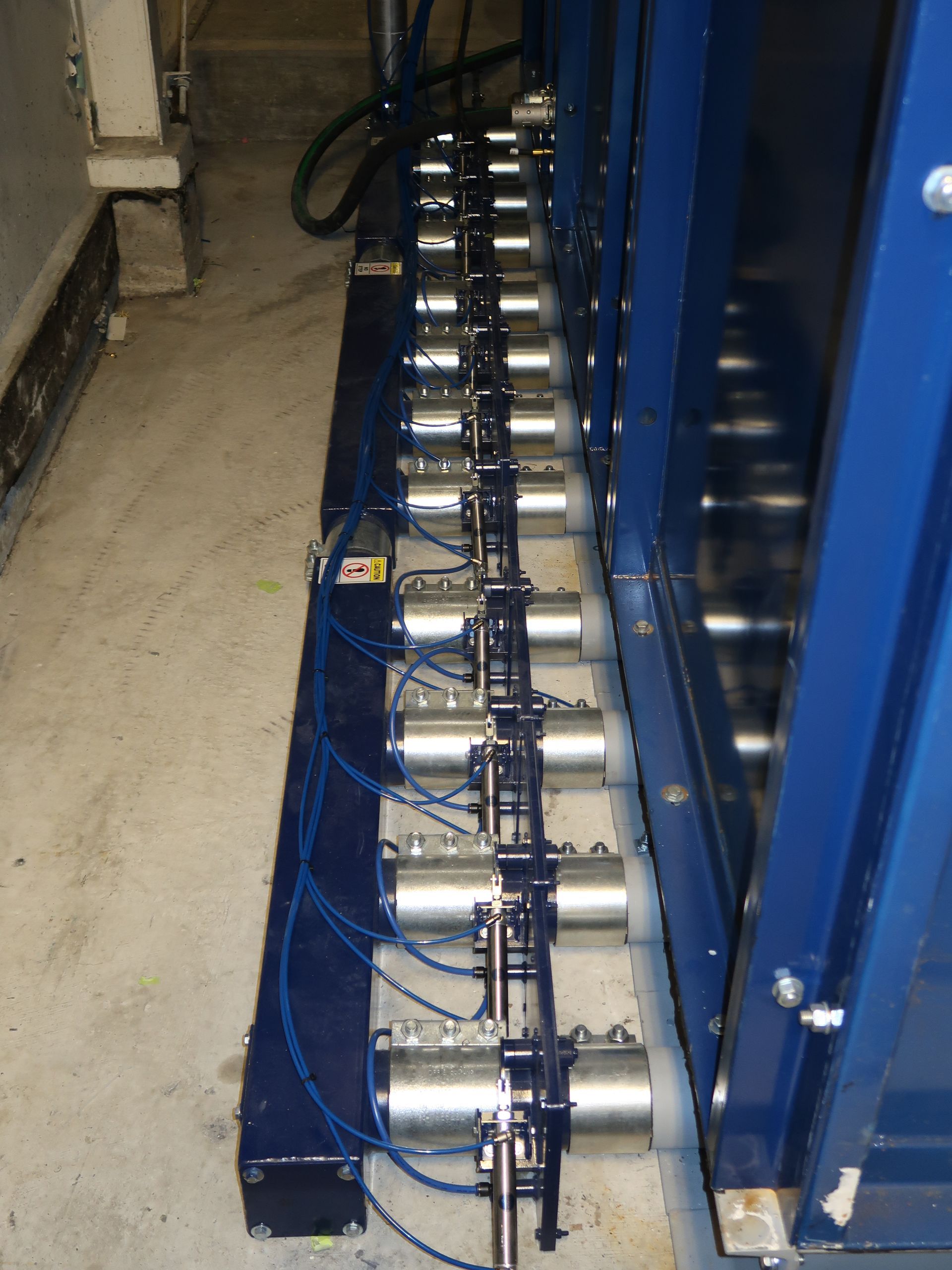A line of silver pneumatic cylinders on a blue metal frame, installed next to a large blue machine in a concrete room.