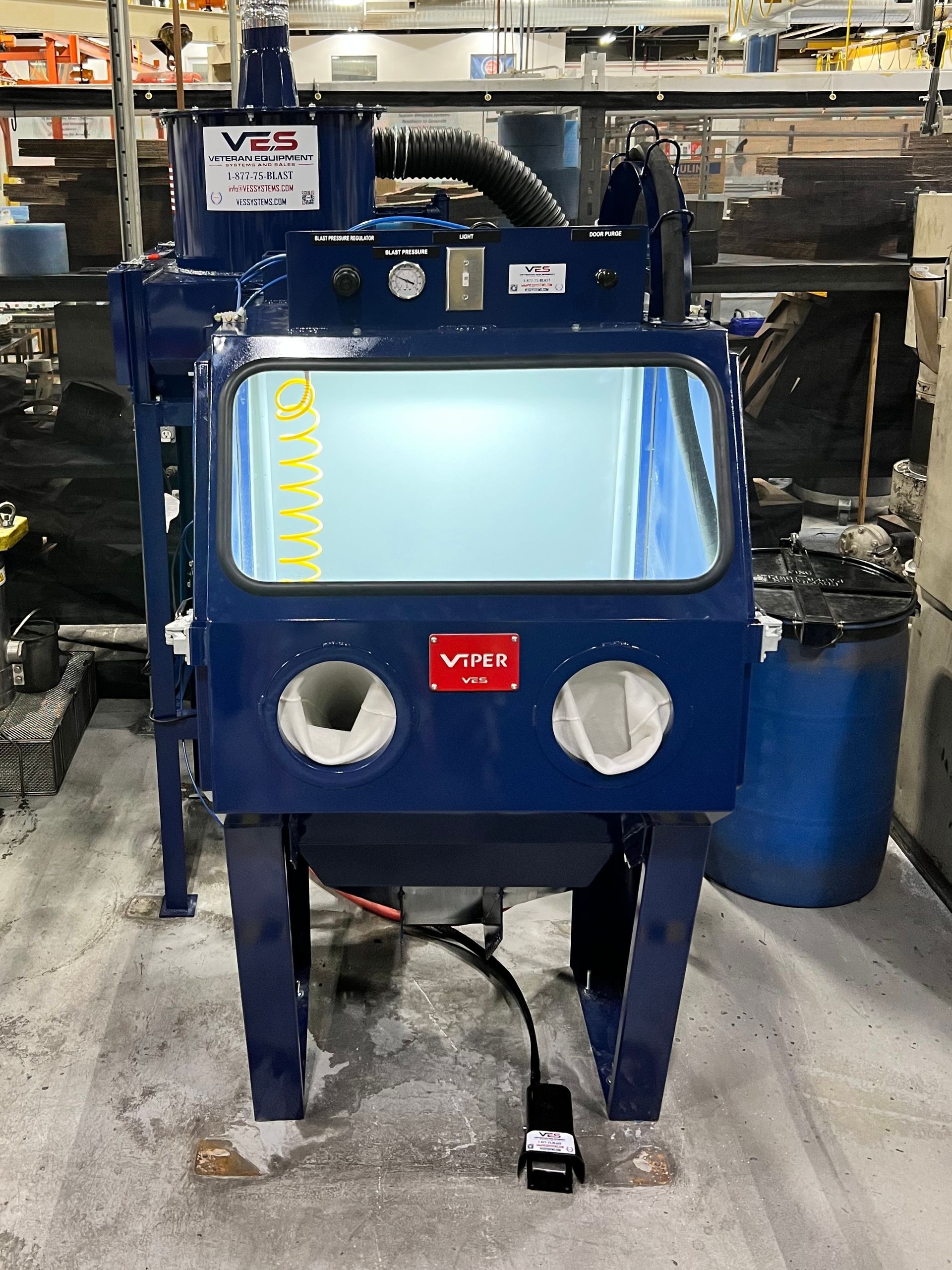 Blue sandblasting cabinet with clear viewing window and armholes, on a factory floor.